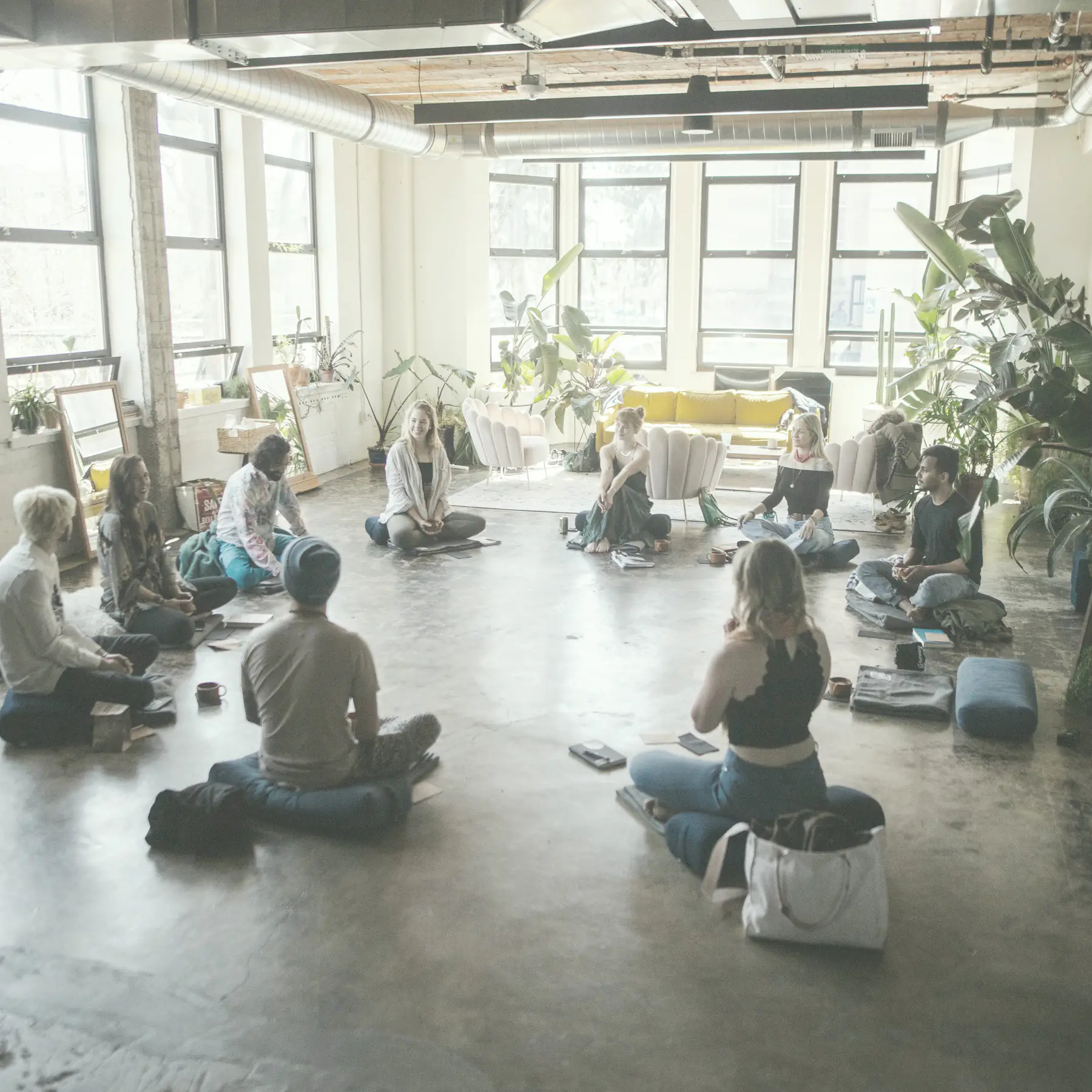 A circle of people sitting on the floor of a bright loft, meditating together.